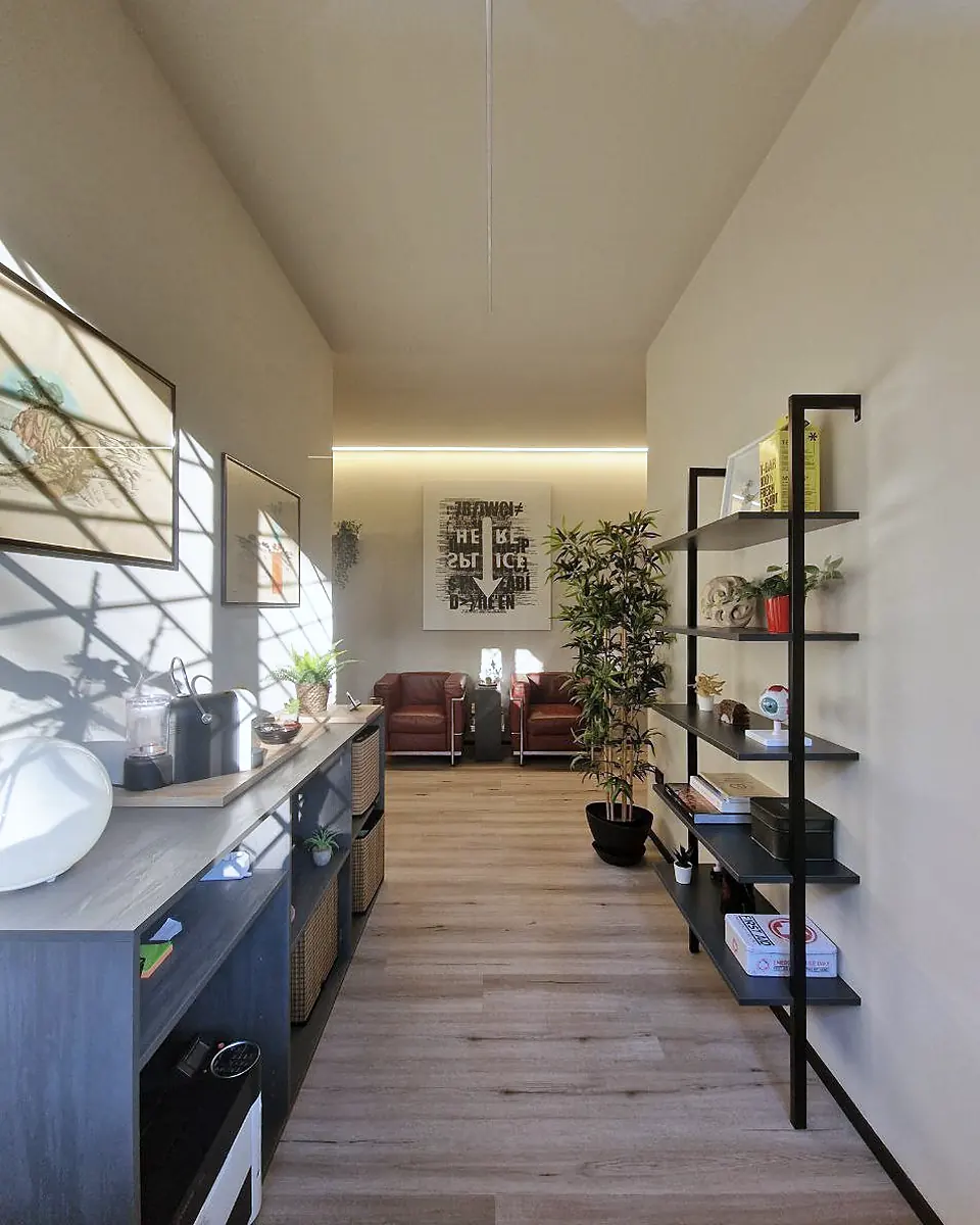 L'ingresso di Via Sismonda: un corridoio luminoso con scaffali, piante e la macchina del caffè; in fondo le poltrone della sala d'attesa.